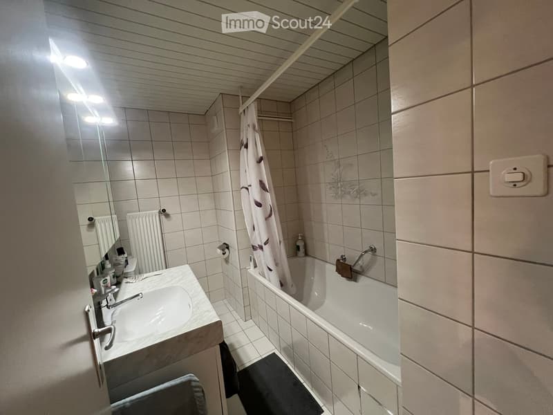 A modern bathroom with a white tiled wall, a sink with a faucet, a bathtub with a shower curtain, a mirror, a radiator, and a towel on the floor.