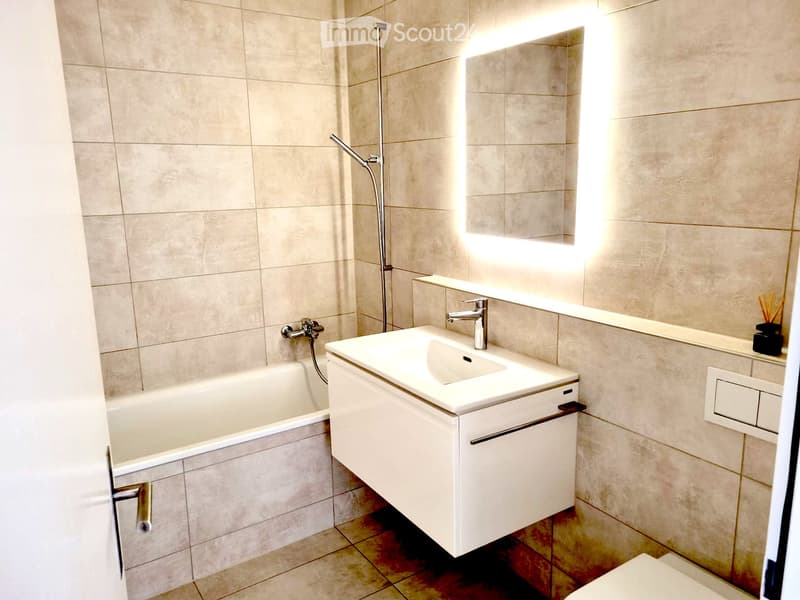 Modern bathroom with beige tiled walls, a white bathtub, a white sink with a chrome faucet, a large mirror, and a toilet.