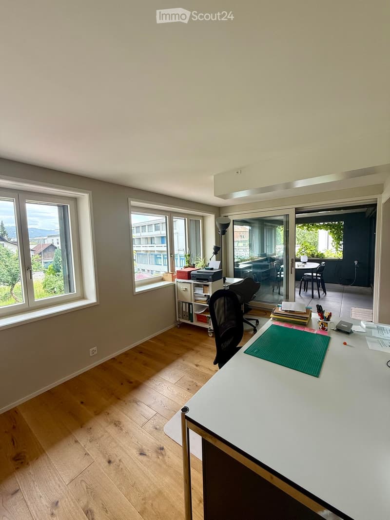 Office space with wooden flooring, large windows, desk, chair, lamp, printer, shelf, and a view of the balcony area with additional furniture.