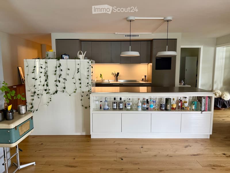 Modern kitchen with grey cabinets, white island, pendant lights, and a variety of bottles on the counter. Wooden floor and decorative plants visible.