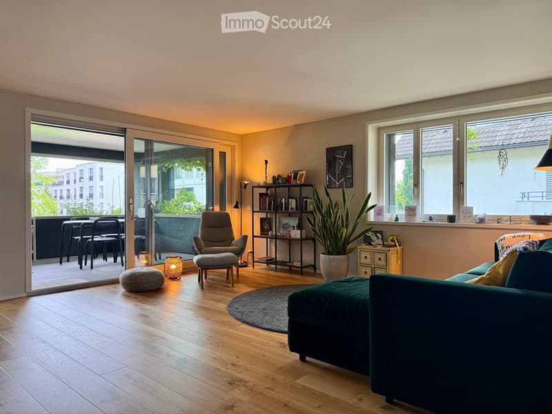 Modern living room with wooden flooring, large windows, and a balcony. Features a green sofa, armchair, small table, shelves, and a potted plant.