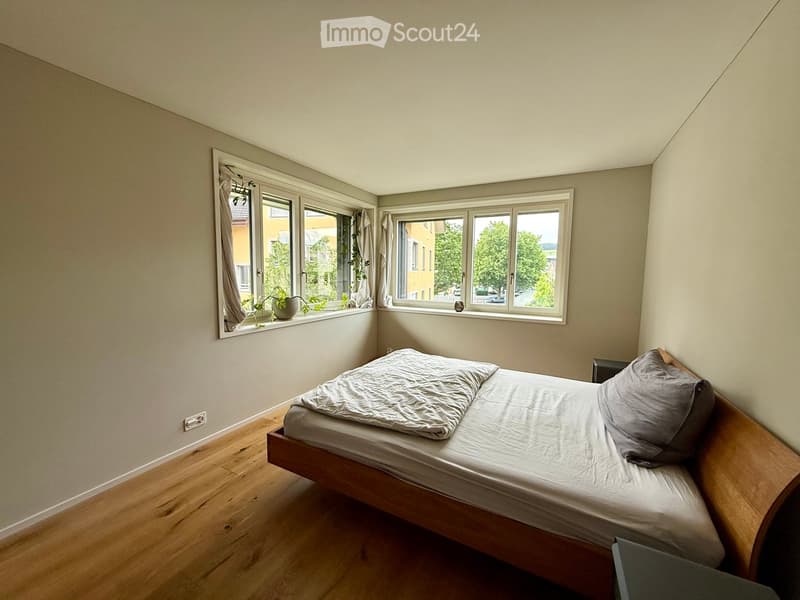 Cozy bedroom with wooden floor, large windows, and a neatly made bed with pillows and a duvet.