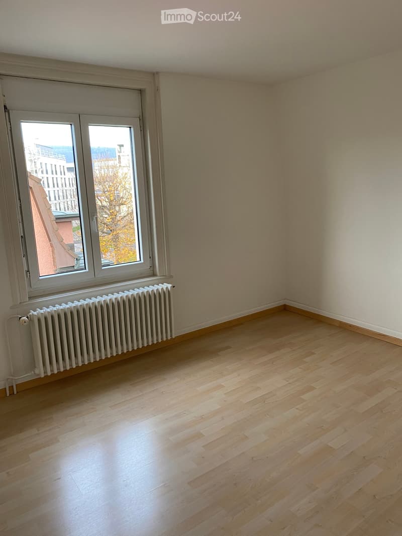 Empty room with wooden flooring, white walls, and a large window with a radiator below it.