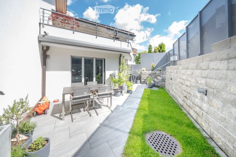 Balcony with table and chairs, potted plants, green lawn, stone wall, and staircase.