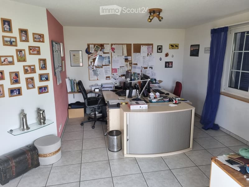 Office space with a desk, chair, computer setup, and various office supplies. The room features a bulletin board, framed pictures, and a glass shelf with decorative items.