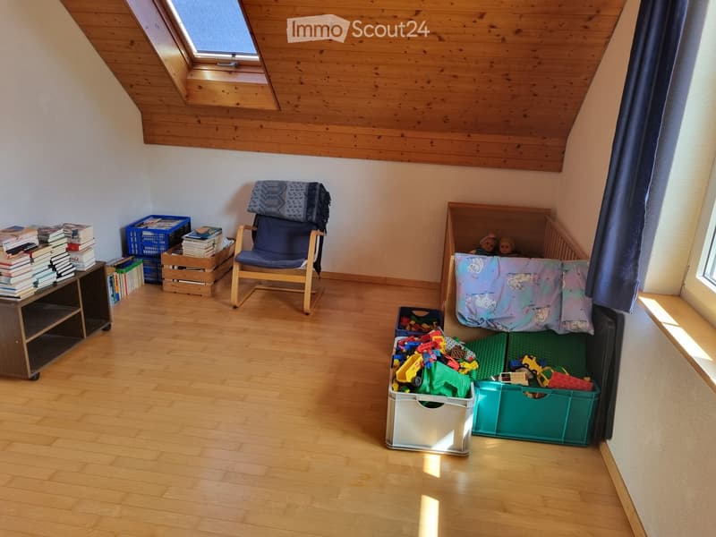A cozy, well-lit room with wooden flooring, a slanted wooden ceiling, and a skylight window. Features include a comfortable chair with a cushion, a bookshelf filled with books, a bed with a colorful blanket and pillows, and several toy boxes filled with t