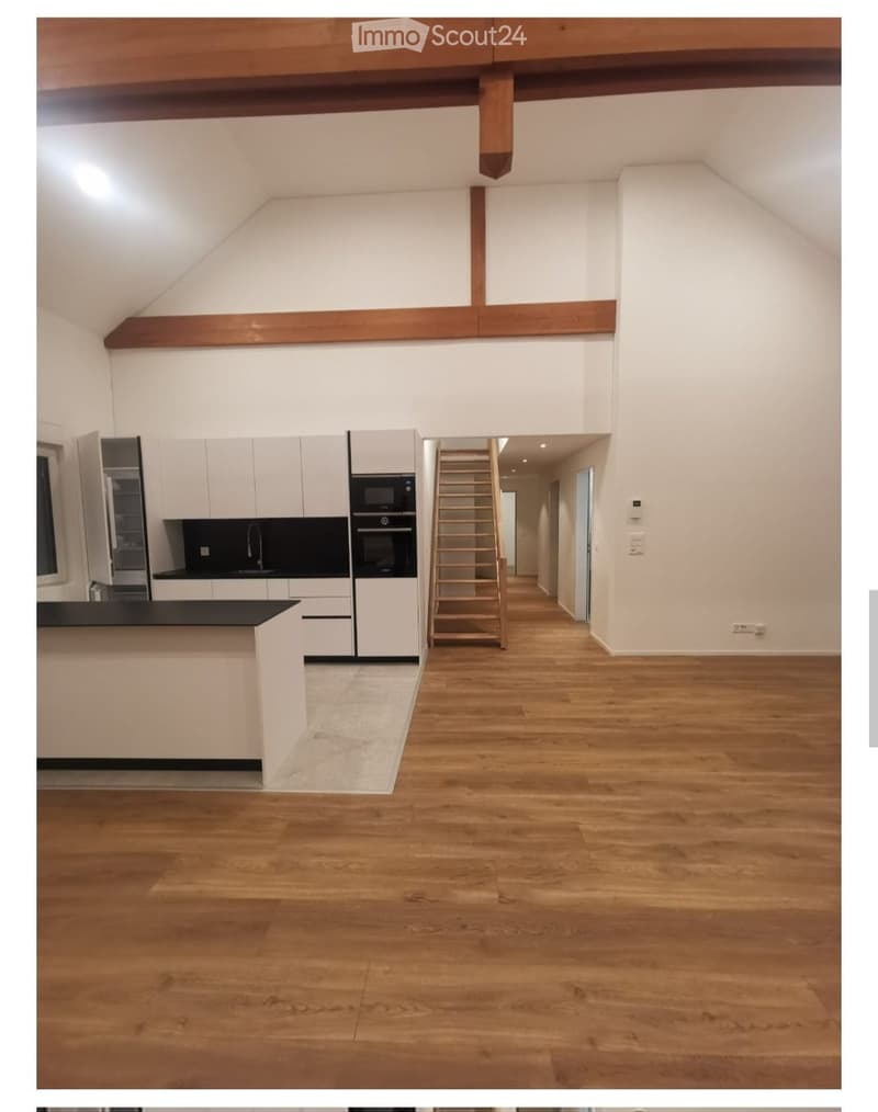 Modern kitchen area with white cabinets, stainless steel appliances, and a wooden staircase leading to an upper level. The space features wooden flooring and exposed wooden beams on the ceiling.
