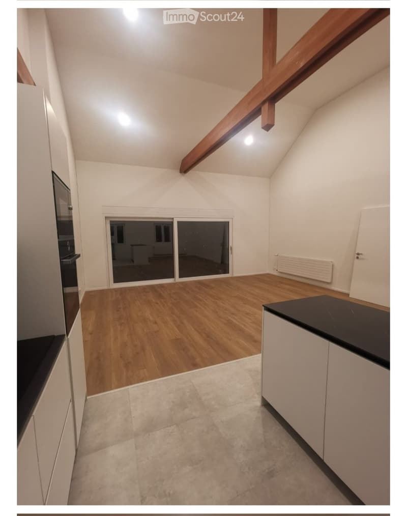 An empty room with wooden flooring, white walls, and a large sliding glass door leading to another space. The room features a white cabinet and a radiator on one wall.