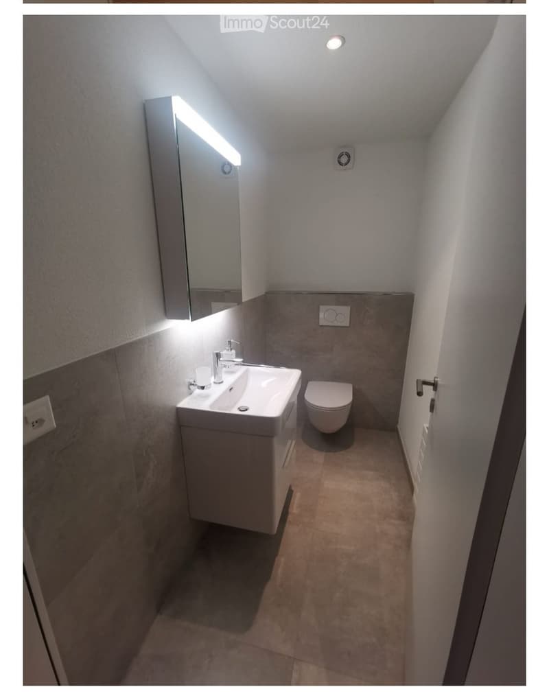Modern bathroom with a white sink, toilet, and illuminated mirror. The walls are tiled in a neutral color scheme, and the floor is tiled with a warm tone.