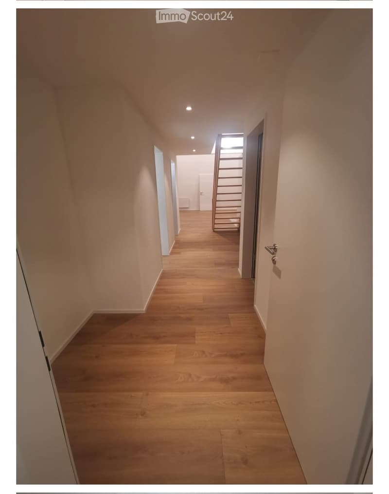 A clean, well-lit hallway with light wooden flooring and white walls. The corridor features recessed ceiling lights and doors leading to other rooms.