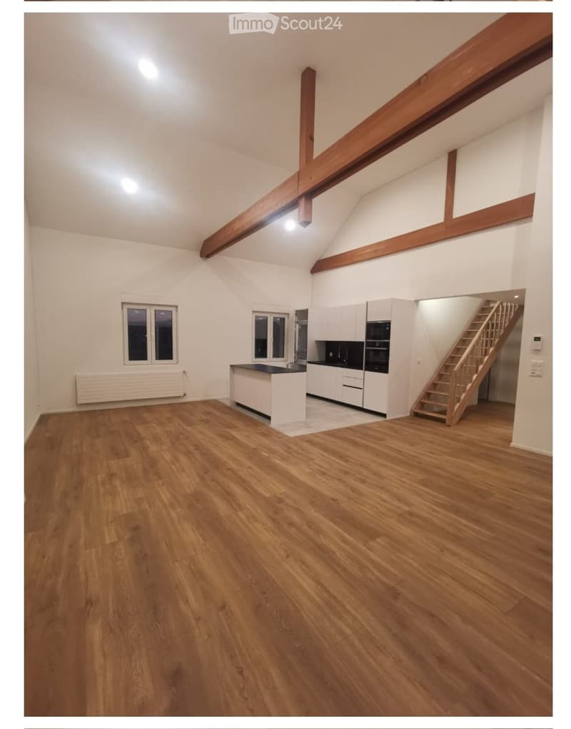 Empty room with wooden flooring, white walls, exposed wooden beams, and a staircase leading to another level. The room features a small kitchen area with cabinets and appliances visible in the background.