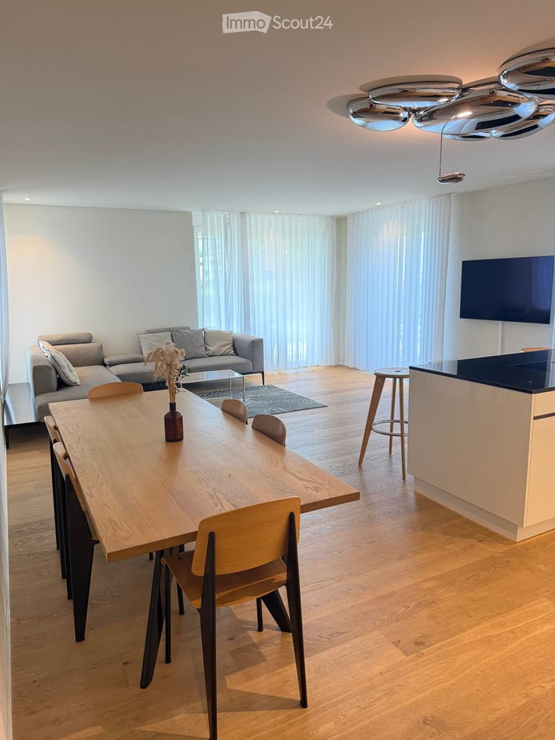 Modern open-plan living and dining area with a grey sofa, wooden dining table, chairs, a kitchen counter, and a television mounted on the wall.