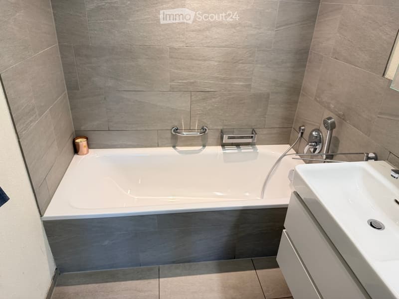 Modern bathroom with a bathtub, sink, and chrome fixtures, featuring grey tiled walls and a small candle on the edge of the tub.