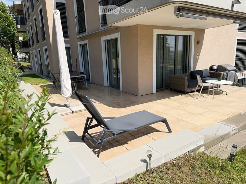Modern outdoor patio with tiled flooring, lounge chair, sofa, small table, and umbrella. Adjacent to a building with large glass doors and balconies.