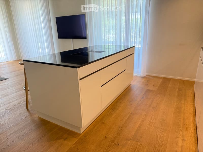 Modern kitchen with a white island featuring a glass countertop and a mounted screen above it. The room has wooden flooring and large windows with vertical blinds.