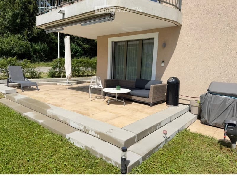 Balcony area with a sofa, coffee table, chairs, umbrella, and lounge chair, connected to a house with a covered walkway.