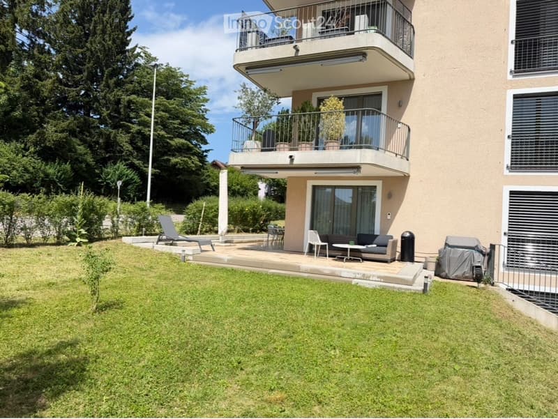 Well-maintained garden with a raised patio area, featuring a sofa, table, chairs, and a lounge chair. The patio is connected to a modern building with balconies and potted plants.