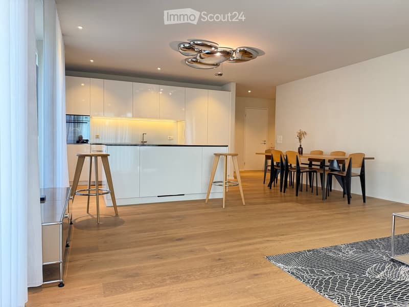 Open-plan living and dining area with modern white cabinets, wooden flooring, and a sleek chandelier. Includes a dining table with chairs, bar stools, and a vase on the table.