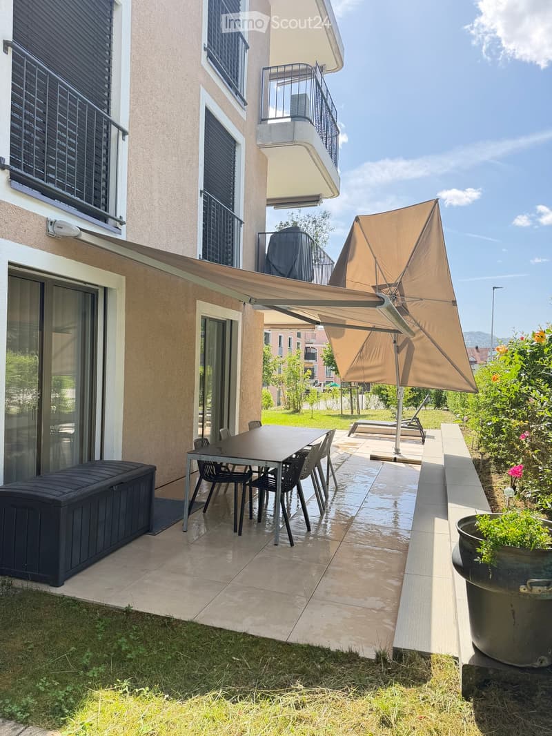 Balcony area with a table, chairs, umbrella, storage box, and flower pot, adjacent to a building with large windows and a covered walkway.