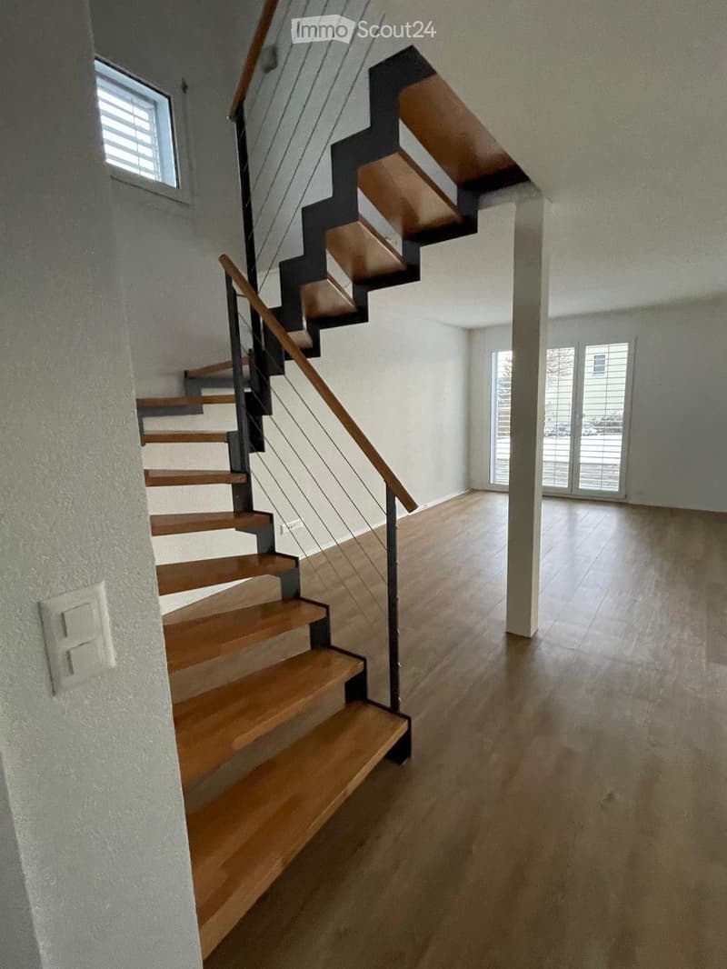 Modern hallway with wooden staircase, light-colored walls, and large windows at the end.