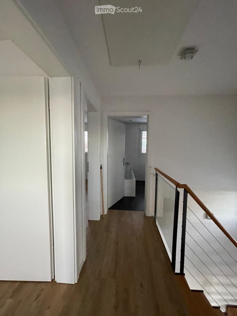 A narrow hallway with wooden flooring, white walls, and a staircase leading upwards. A small bathroom is visible at the end of the hallway.