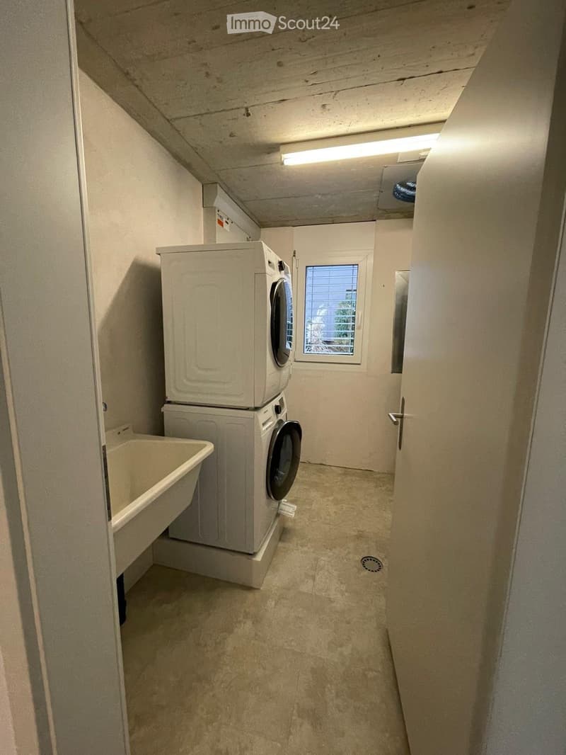 Laundry area with stacked washing machine and dryer, a bathtub, and a window with blinds.