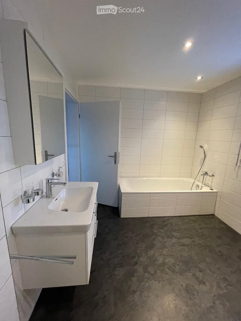 Modern bathroom with white tiled walls, a white sink with a chrome faucet, a bathtub, and a large mirror above the sink. The floor is dark grey.