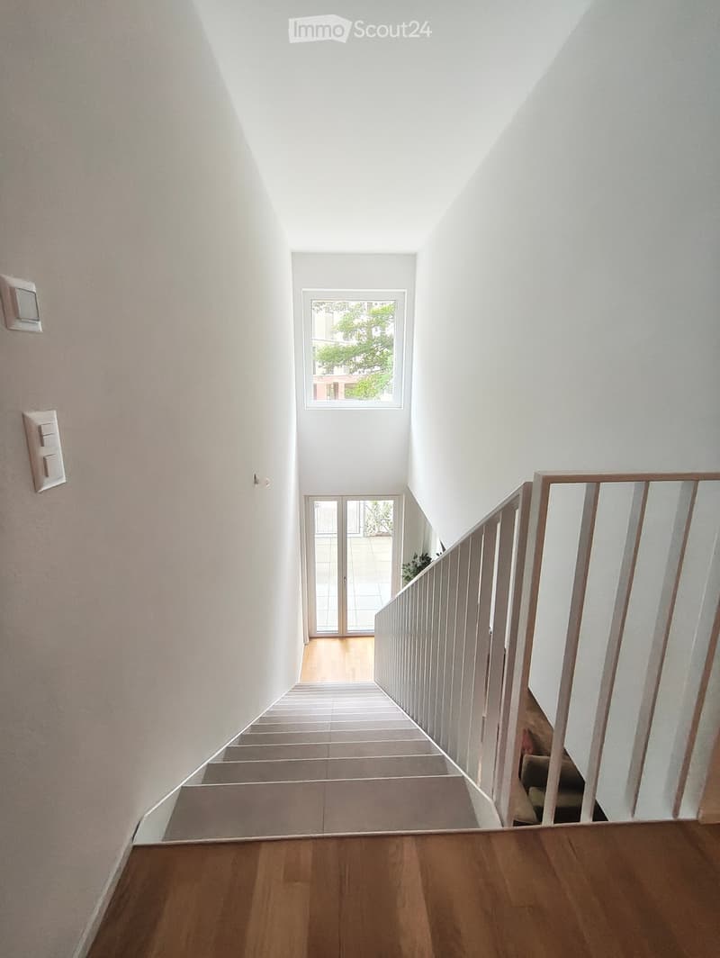 Modern hallway with wooden flooring, white walls, and a staircase leading upwards. Features a window at the top of the stairs and a glass door leading to an outdoor area.