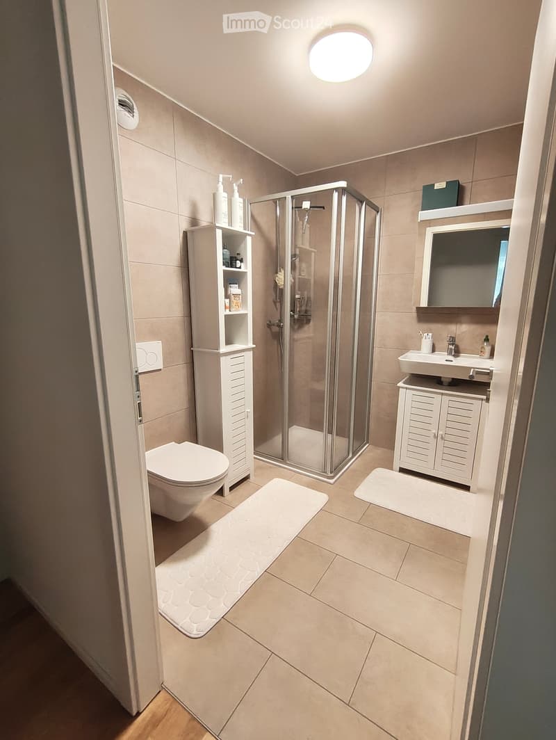 Modern bathroom with beige tiles, a toilet, a sink with a mirror, a glass-enclosed shower, and storage cabinets.