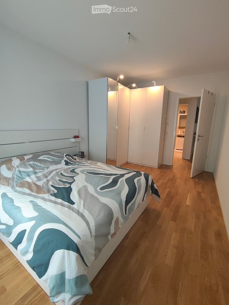 Modern bedroom with a large bed covered in a blue and white abstract duvet, a built-in wardrobe, and a nightstand with a lamp. The room has wooden flooring and white walls.