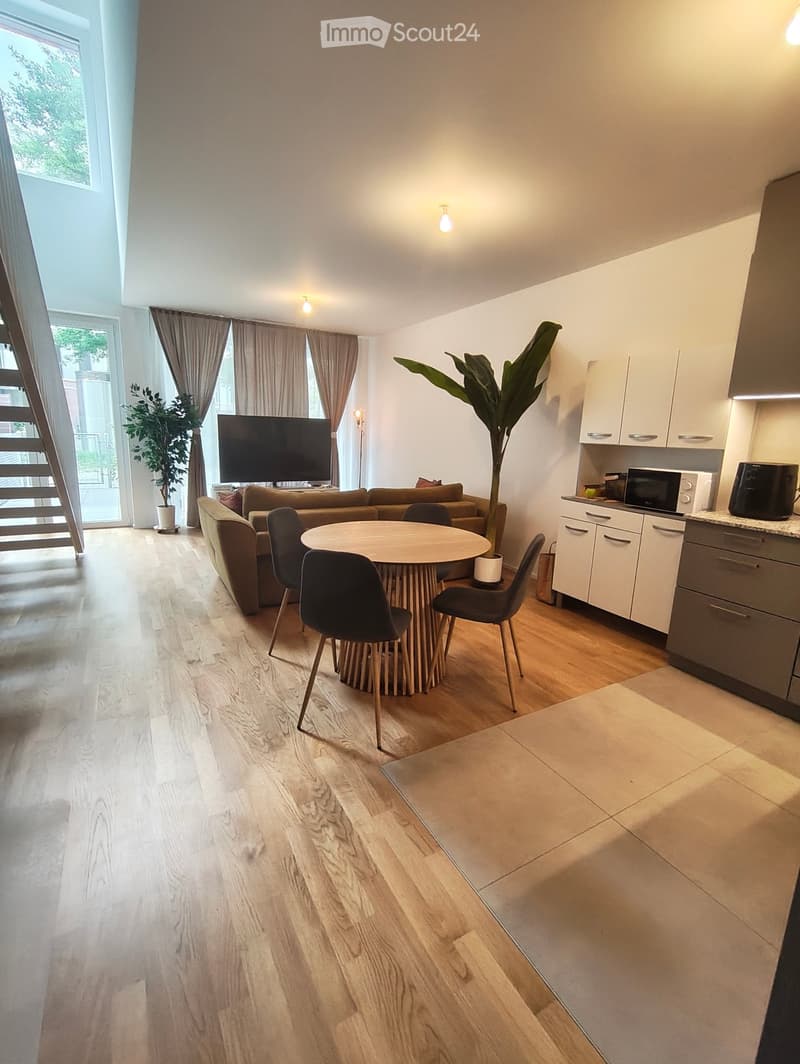 Open-plan living and dining area with a sofa, dining table, chairs, and a television. Kitchen area features white cabinets and a microwave. Wooden flooring throughout.