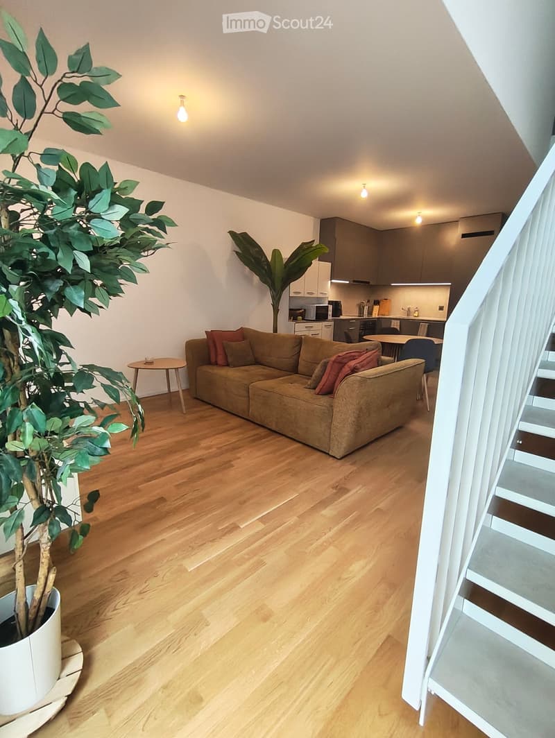 Open-plan living and dining area with wooden flooring, a large sofa, a small round table, a potted plant, and a modern kitchen in the background.