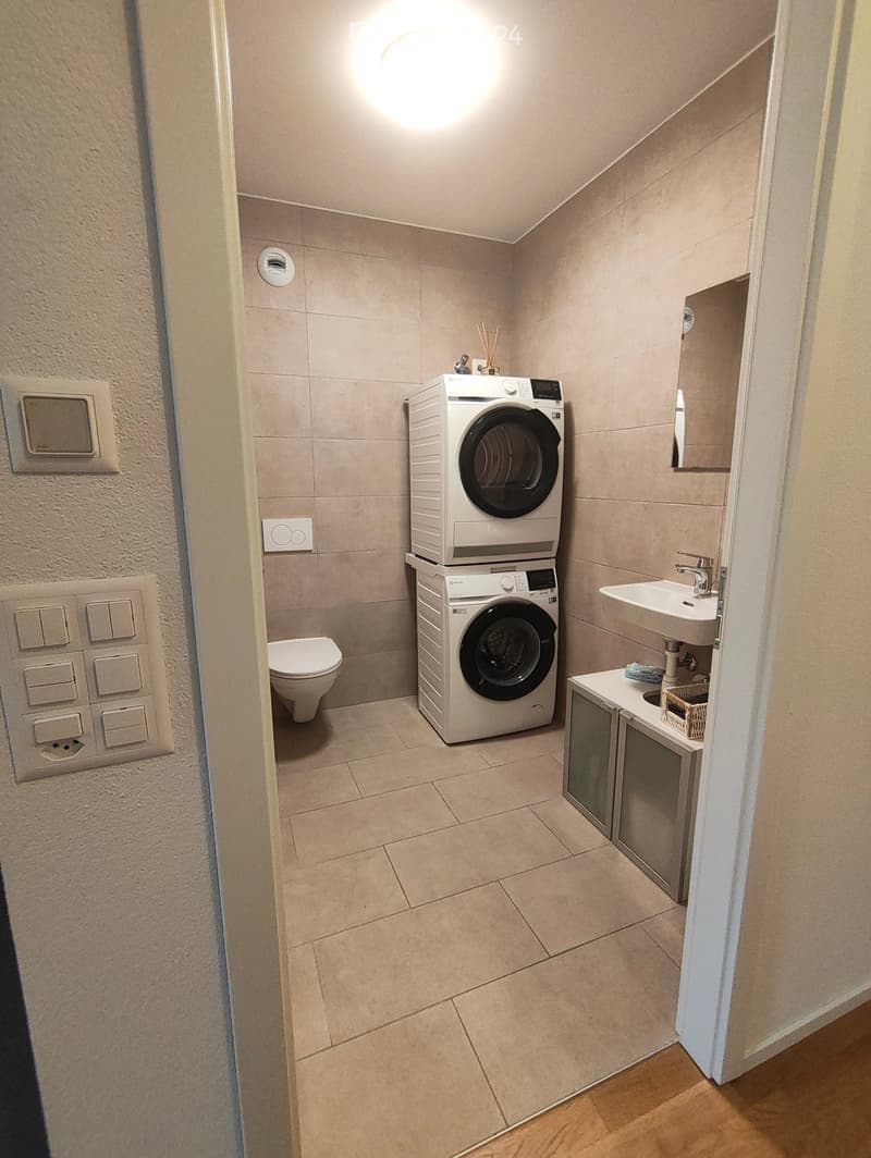 Compact bathroom with a toilet, stacked washing machine and dryer, sink with mirror, and small cabinet.