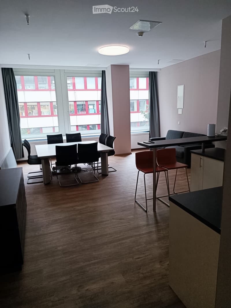 Open-plan room with wooden flooring, large windows, a sofa, a dining table with chairs, and a bar area with stools.
