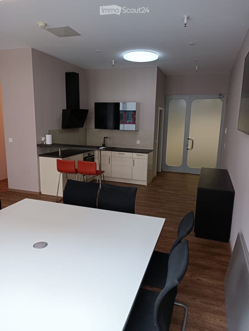 Open-plan space with a kitchen area and a dining area. The kitchen features white cabinets, a black countertop, a sink, a faucet, an oven, and a range hood. A screen is mounted on the wall. The dining area includes a white table with four black chairs and