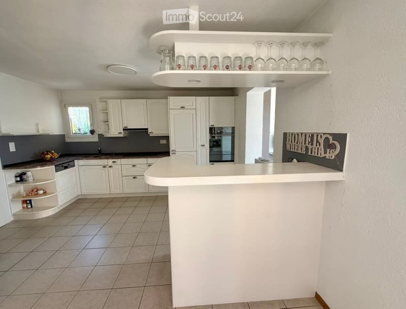 Modern kitchen with white cabinets, a bar counter, and a refrigerator. Shelves hold cups and a bowl of fruit is on the counter.