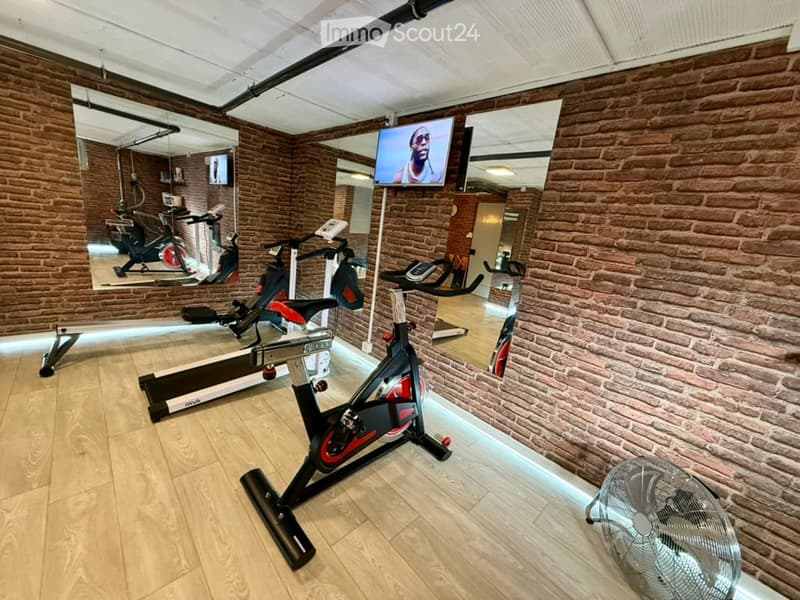 Fitness room with wooden flooring, brick walls, exercise bike, elliptical machine, fan, and a screen mounted on the wall.