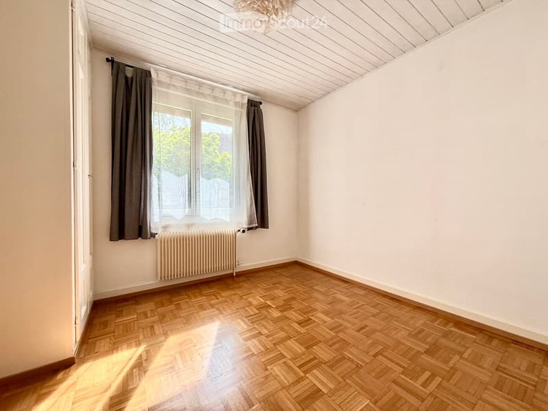 Empty room with wooden parquet flooring, white walls, large window with curtains, radiator, and a light fixture on the ceiling.