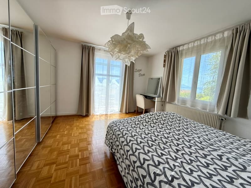 Bright bedroom with wooden flooring, large windows offering a view, a bed with patterned bedding, a wardrobe, a desk with a chair, and a decorative hanging lamp.
