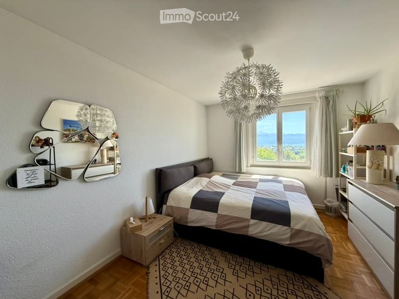 Modern bedroom with wooden flooring, large window offering a scenic view, bed with checkered duvet, nightstand, mirror, and decorative shelf.