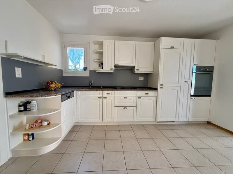 Modern kitchen with white cabinets, granite countertops, tiled floor, and built-in appliances including an oven and dishwasher. A fruit bowl and small shelves with jars are visible on the counter.