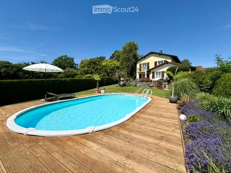 A wooden deck surrounds a swimming pool, with lounge chairs, an umbrella, and a house in the background.