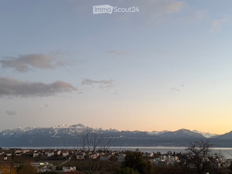 Panoramic view of a town with houses, surrounded by mountains and a body of water under a partly cloudy sky.