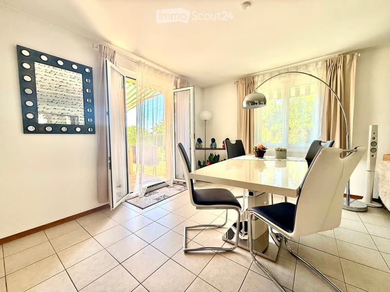 Modern dining room with a white dining table, black chairs, a large mirror on the wall, and a floor lamp. The room features large windows with sheer curtains and a sliding glass door leading to a balcony.