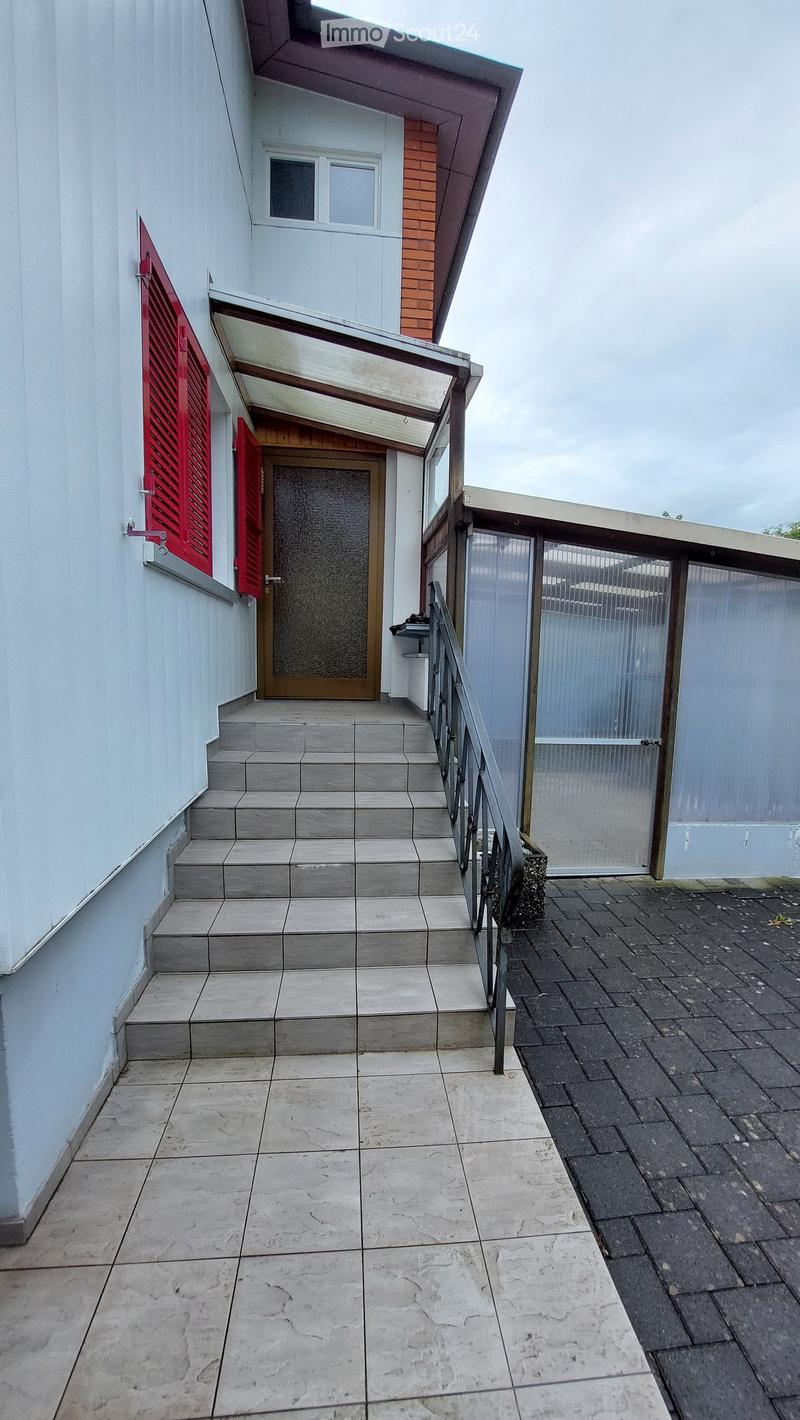 Outdoor area with tiled flooring, steps leading up to a wooden door, metal railing, and a small covered entrance area.