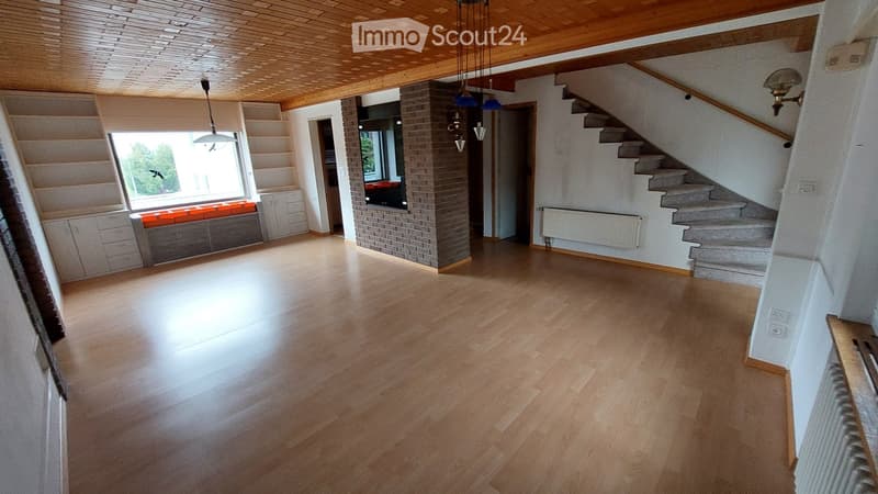 Empty room with wooden flooring, white walls, staircase leading upwards, and a window with a bench seat.