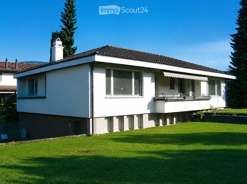 Two-story house with white exterior, dark tiled roof, multiple windows, and a covered balcony area. The house is surrounded by a well-maintained lawn and trees.