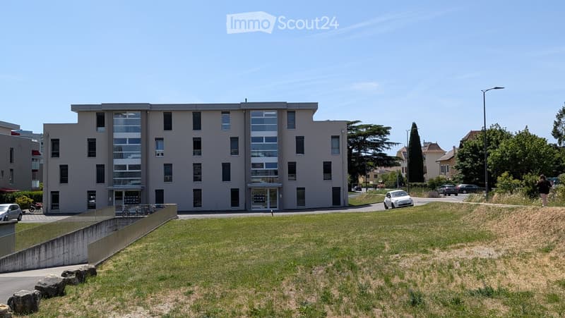 Modern multi-story building with large windows and a raised ground floor, surrounded by a grassy area and parked cars.