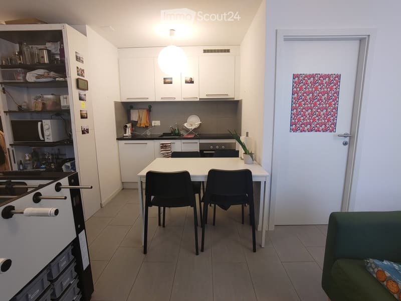 Modern kitchen and living area with white cabinets, a dining table with chairs, a sofa, and a potted plant. The kitchen features a refrigerator, microwave, and oven, with a small seating area visible.
