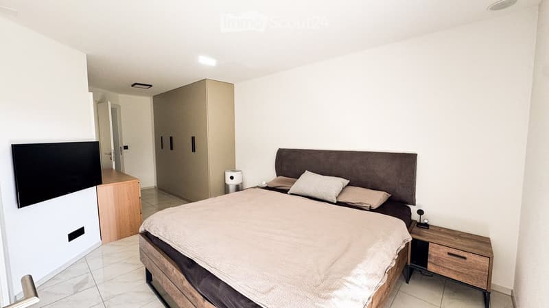 Modern bedroom with a large bed, headboard, nightstand, wardrobe, and a screen mounted on the wall.